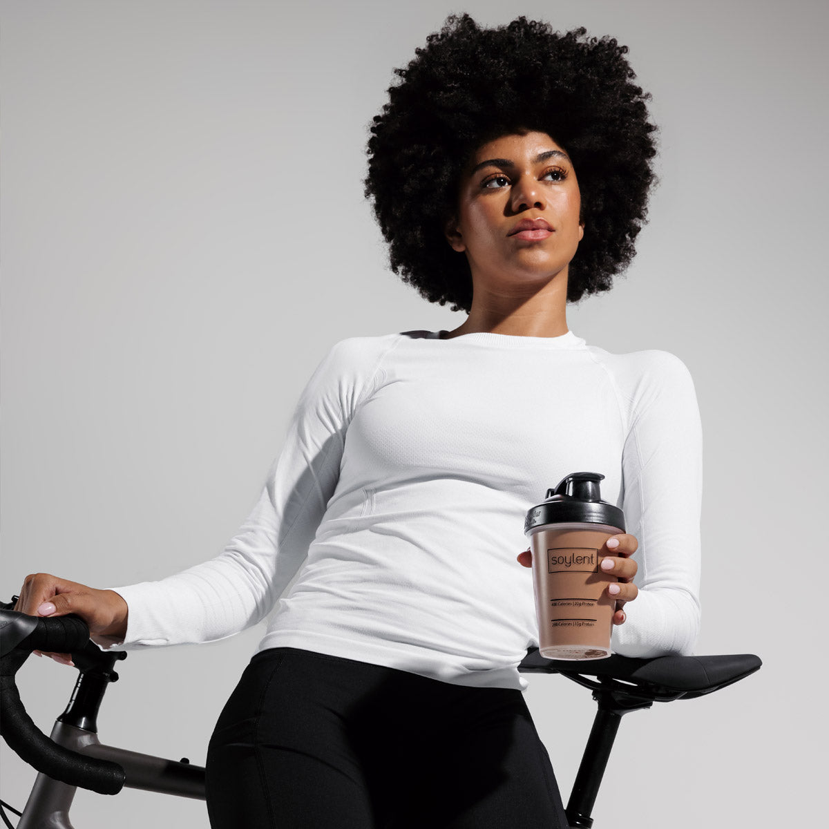 Woman holding chocolate Soylent Complete Protein Powder while standing next to bike.